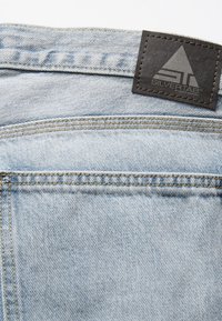 Tejido de mezclilla azul claro con textura de tejido visible, con una etiqueta de cuero negro embosada con "SILVERTAB" y un logo triangular.