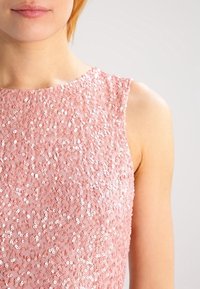 rosa paljetterad topp med rundad halsringning och ärmlös design, med en strukturerad yta skapad av överlappande glänsande paljetter.