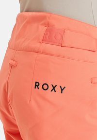 Koraalroze Roxy-broek met een gladde textuur, voorzien van een label met de merknaam in zwart gedrukt en één achterzak met een subtiele stikdetail.