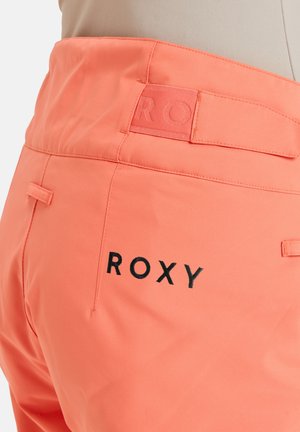 Koralle Roxy-Hose mit glatter Textur, versehen mit einem Etikett mit dem eingeprägten Markennamen in Schwarz und einer hinteren Tasche mit feinen Nahtdetails.