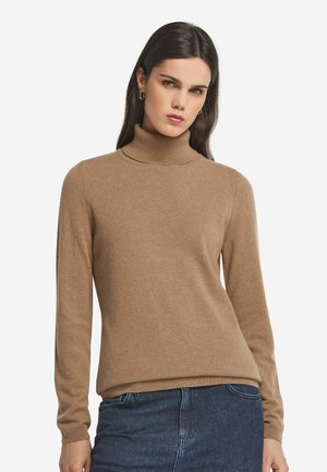 Kvinde med langt mørkt hår iført guldringøreringe, en brun højhalset sweater og blå jeans, stående mod en hvid baggrund.