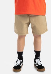 Pantalones cortos caqui con un corte recto, dobladillo doblado y bolsillos frontales. Zapatillas negras con detalles en blanco y calcetines de rayas negras. Camisa naranja arriba.