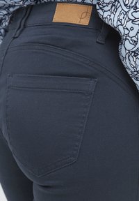 Tmavomodré džíny z denimu s hladkou textúrou, s jednou zadnou kapsou a hnedou koženou nášivkou na páse. Viditeľné jemné detaily šitia.