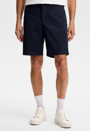 Mann trägt marineblaue Shorts, weiße Socken und weiße Turnschuhe, steht vor einem schlichten hellen Hintergrund.
