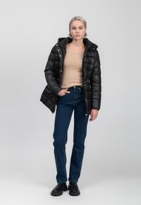 Schwarze Steppjacke mit Kapuze, goldenen Beschlägen und einem gesteppten Muster, getragen über einem beigen Oberteil und blauen Jeans, mit schwarzen Ankle-Boots.