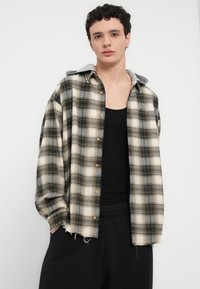 Καρό πουκάμισο σε μπεζ, γκρι και μαύρο, με γκρι κουκούλα, κουμπιά μπροστά και ξεφτισμένο τελείωμα. Φορεμένο πάνω από μαύρο τοπ και σκούρο παντελόνι.