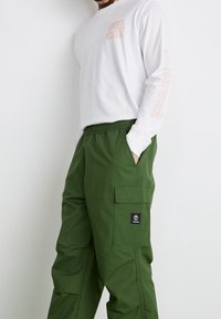 Vit långärmad skjorta med orange grafik på framsidan och ärmarna, i kombination med gröna cargobyxor med sidofickor och en logotypapplikation.