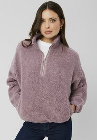 Pull en polaire violet avec un col zippé quart, coupe décontractée, surface texturée et poignets élastiqués. Porté sur une chemise blanche.