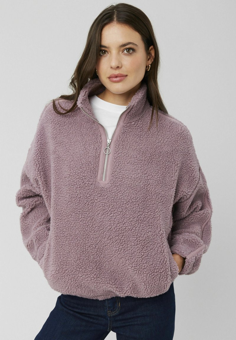 Pull en polaire violet avec un col zippé quart, coupe décontractée, surface texturée et poignets élastiqués. Porté sur une chemise blanche.