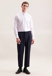 Camicia bianca a maniche lunghe con bottoni abbinata a pantaloni plissettati blu navy e scarpe Oxford nere. Design semplice e vestibilità su misura.