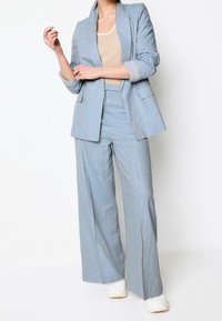 Blazer bleu clair sur mesure et pantalon large assorti, confectionnés en tissu texturé. Associés à un haut beige et des baskets blanches.