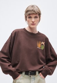 Καφέ φούτερ με φαρδιά εφαρμογή, ριμπ μανσέτες και λαιμόκοψη. Διαθέτει γραφικά μοτίβα σε χρυσό, πράσινο και εκρού στο μπροστινό μέρος.