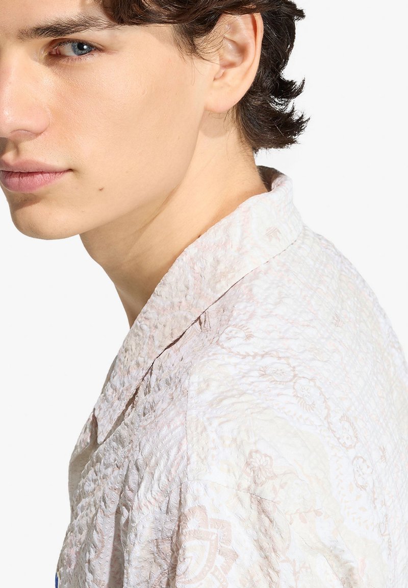 Giovane uomo dalla pelle chiara e occhi azzurri che indossa una camicia bianca strutturata con un delicato motivo floreale, che guarda leggermente di lato.