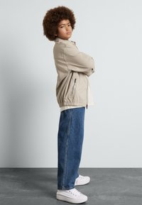 Beige, översize jacka med elastisk midja och sidofickor; i kombination med lösa blå jeans och vita sneakers. Personen står med armarna i kors.