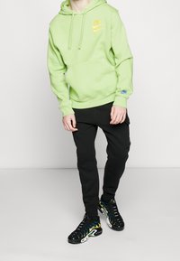 Ljusgrön hoodie med en gul Nike-logotyp, kombinerad med svarta joggers och multikolorade sportskor med ett vågigt mönster.