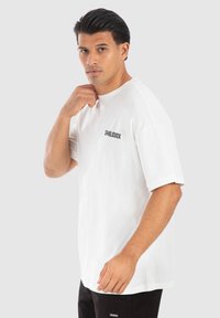 Hvid bomulds t-shirt med korte ærmer, rund halsudskæring og sort logo tekst på venstre bryst. Model poserer foran en neutral baggrund.