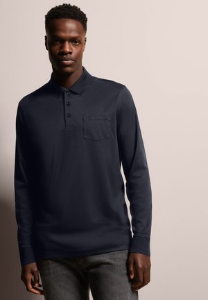 Navyblaues Langarm-Polohemd mit Kragen, Knopfdetails und einer Brusttasche. Glatte Textur und lässige Passform. Getragen mit grauen Jeans.