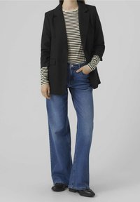 Blazer noir ajusté par-dessus une chemise à rayures noires et blanches à manches longues, associé à un jean bleu à jambe large et des chaussures plates noires.