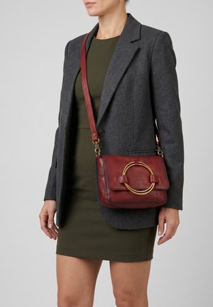 Vrouw draagt een donkergrijs blazer en een olijfgroene jurk met een rode leren crossbody tas met een groot gouden cirkelvormig detail.