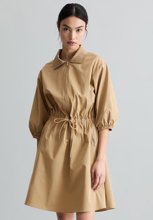 Jeune femme aux cheveux foncés portant une robe beige zippée avec des manches bouffantes et une taille à cordon, debout devant un fond uni.