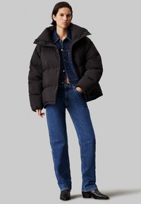 Zwarte pufferjack met een hoge kraag, gedragen over een spijkerjack. Blauwe rechte jeans en zwarte enkellaarsjes. Soepele texturen, casual ontwerp.