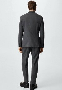 Costume gris à coupe ajustée, comprenant une veste structurée et un pantalon slim. Tissu à texture douce, avec des détails de couture subtils. Bottes noires.