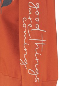 Oranges Sweatshirt mit weißem, geschwungenem Text "Gute Dinge kommen" entlang des Ärmels. Weiches Material mit lockerer Passform.