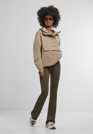 Naise, kellel on afro soeng, pruuni tuunika, oliivrohelised laiade säärega püksid, valged vabaajajalanõud ja päikeseprillid, seisab ühetaustalise heleda tausta ees.