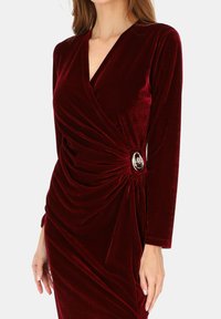 Długa sukienka z długim rękawem z aksamitu w głębokim bordowym kolorze, z kopertowym fasonem, marszczeniem po bokach oraz srebrnym kwiatowym akcentem w talii. Miękka w dotyku.