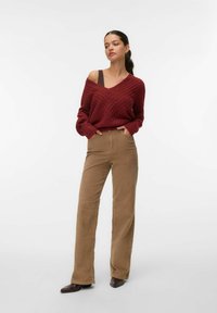 Bordeaux gebreide trui met V-hals en ribstructuur, gecombineerd met khaki corduroy wijde broekspijpen. Bruine schoenen, handen op de heupen, neutrale achtergrond.