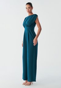 Model poses in a teal jumpsuit with a fitted bodice and wide legs, paired with nude high-heeled sandals against a light background.