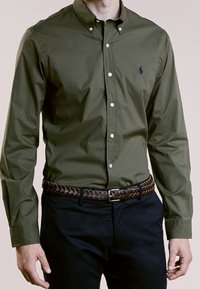 Oliwkowa koszula na guziki z ostrym kołnierzykiem, z białymi guzikami i małym haftowanym logo. Połączona z ciemnymi spodniami i plecionym brązowym paskiem.