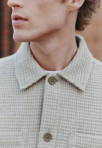 Beige textured jacket with a button-up front, pointy collar, and a patch pocket, featuring two round buttons in an olive hue.