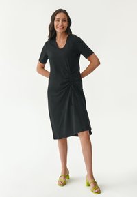 Schwarzes kurzes Kleid mit kurzärmeligen, V-Ausschnitt und seitlichem Raffungselement. Kombiniert mit limonengrünen Blockabsatzsandalen. Glatte Stofftextur.