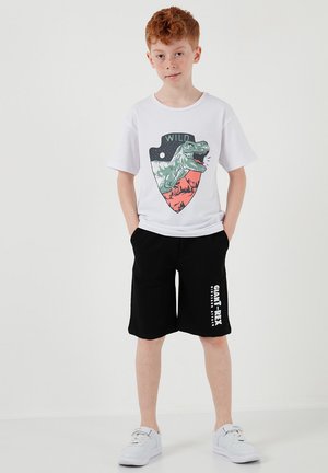 Červenovlasý chlapec v bielom tričku s potlačou dinosaura a čiernych šortkách s bielym textom, stojaci s rukami v vreckách a v bielych teniskách.