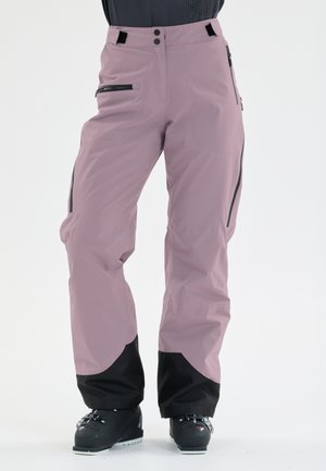Pantaloni da sci viola chiaro con polsini rinforzati neri, tasche con zip, indossati con scarponi da sci neri su una persona in piedi.