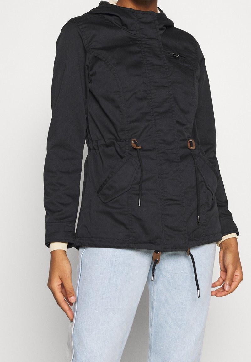 Svart huva jacka med åtdragen midja, med två sidofickor, dragsnören och en texturerad yta. Ljusfärgade ärmslut synliga.