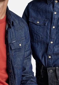 Dos personas llevan chaquetas de mezclilla azul oscuro; una combinada con una camisa roja, ambas con cierres de botones a presión y bolsillos en el pecho.