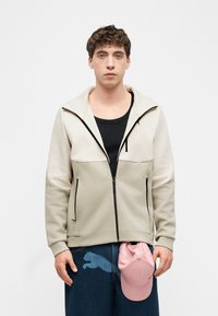 Νεαρός άνδρας που φορά μια μπεζ φούτερ με φερμουάρ πάνω από μια μαύρη μπλούζα, κρατώντας ένα ροζ καπέλο, στέκεται μπροστά σε ένα απλό λευκό φόντο.