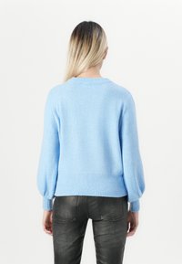 Žena s dlouhými blond vlasy, oblečená v světlém modrém svetru s nabíranými rukávy a černými koženými kalhotami, stojící zády k fotoaparátu na bílém pozadí.