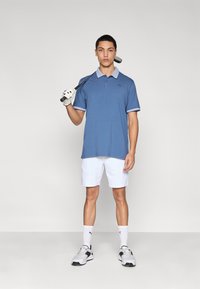 Μπλούζα πόλο σε μπλε χρώμα με αντίθετο ανοιχτό μπλε γιακά, σε συνδυασμό με λευκά σορτς. Το μοντέλο κρατάει ένα μπαστούνι γκολφ, φοράει γάντια και αθλητικά παπούτσια.