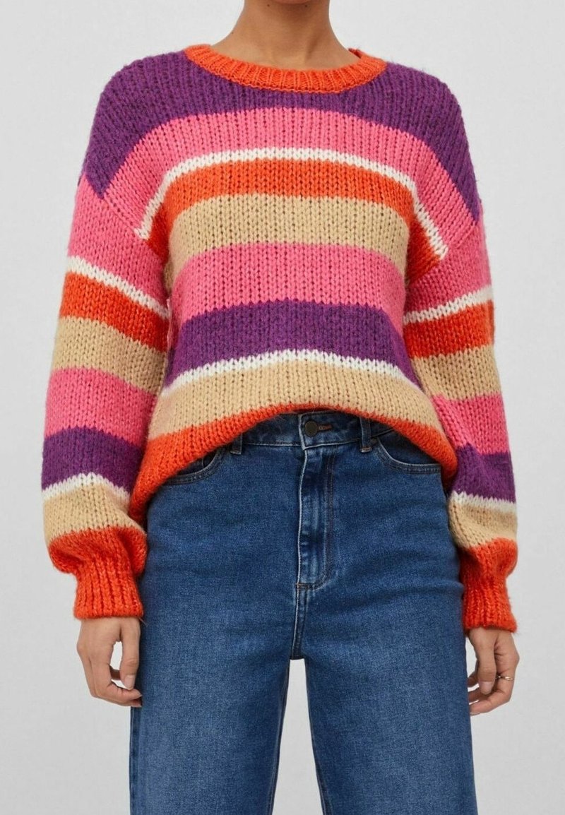 Pull en maille épaisse avec des rayures horizontales orange, violet, rose, beige et blanc, associé à un jean bleu taille haute.