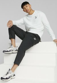 Camisa blanca de manga larga con un logo negro, pantalones ligeros negros y zapatillas grises con detalles negros, sentado en una plataforma blanca.