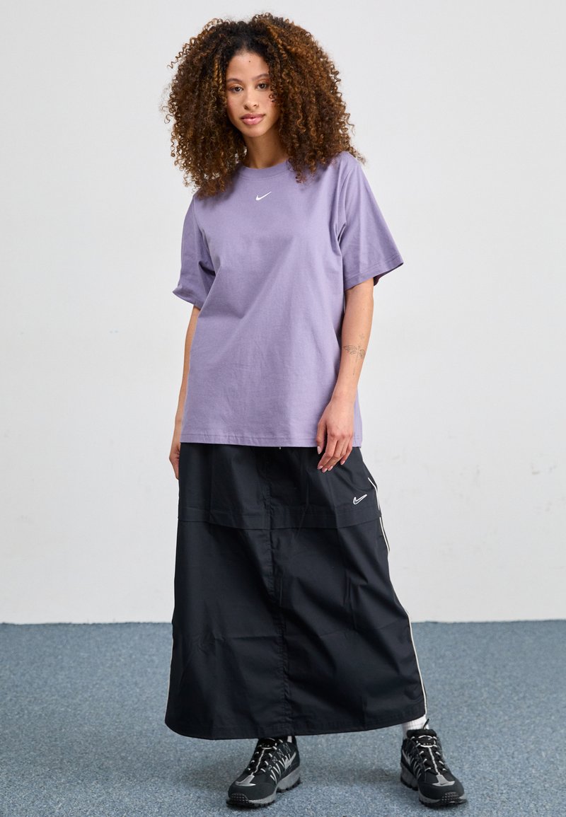 T-shirt en coton violet clair à manches courtes avec un petit logo blanc, assorti à une jupe coupe-vent noire de longueur genoux et des chaussures noires.