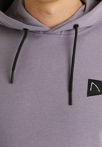 Sudadera con capucha color púrpura claro hecha de tela suave, con una etiqueta negra que lleva un logo blanco. Incluye cordones negros y un ajuste relajado.