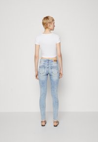 Light blue high-waisted jeans with a faded wash, five pockets, and a small brown logo patch. Worn with a white cropped t-shirt and sandals.