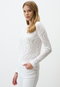 Biały sweter z dzianiny z falistym wzorem, okrągłym dekoltem i długimi rękawami. Noszony z białymi spodniami dla uzyskania monochromatycznego wyglądu. Gładka faktura.