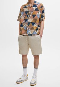 Camisa de manga corta con un patrón de triángulos multicolores en marrón, azul y beige, combinada con pantalones cortos beige. Las zapatillas tienen detalles en blanco y gris.