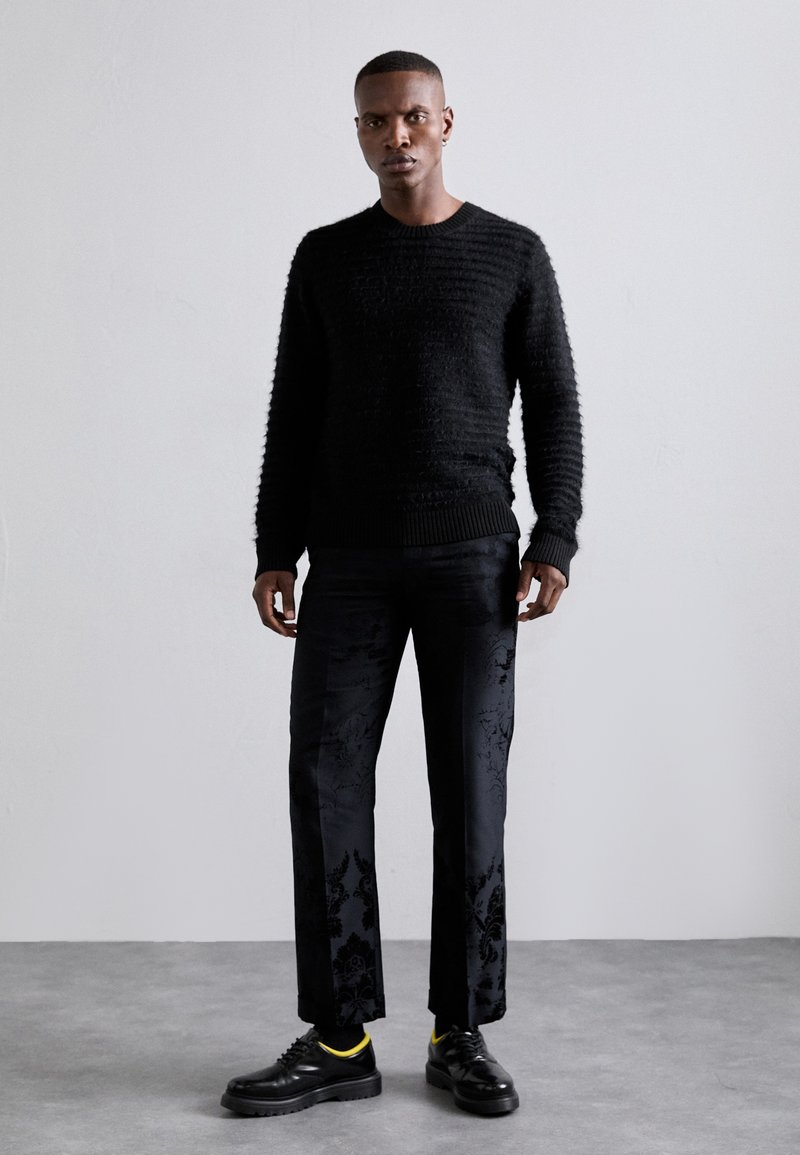 Sort textured sweater med ribbede manchetter, parret med mønstrede sorte bukser. Har skinnende sorte sko med gule accenter. Glat baggrund.
