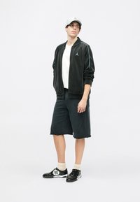 Casaco preto com fecho zip, com detalhes azuis, t-shirt branca, calções escuros e sapatilhas pretas com detalhes brancos e meias de cor clara.